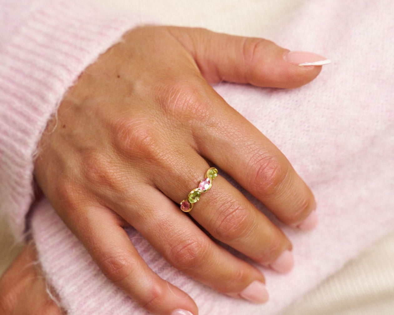 Hand wearing a ring with colorful gemstones on a light pink background