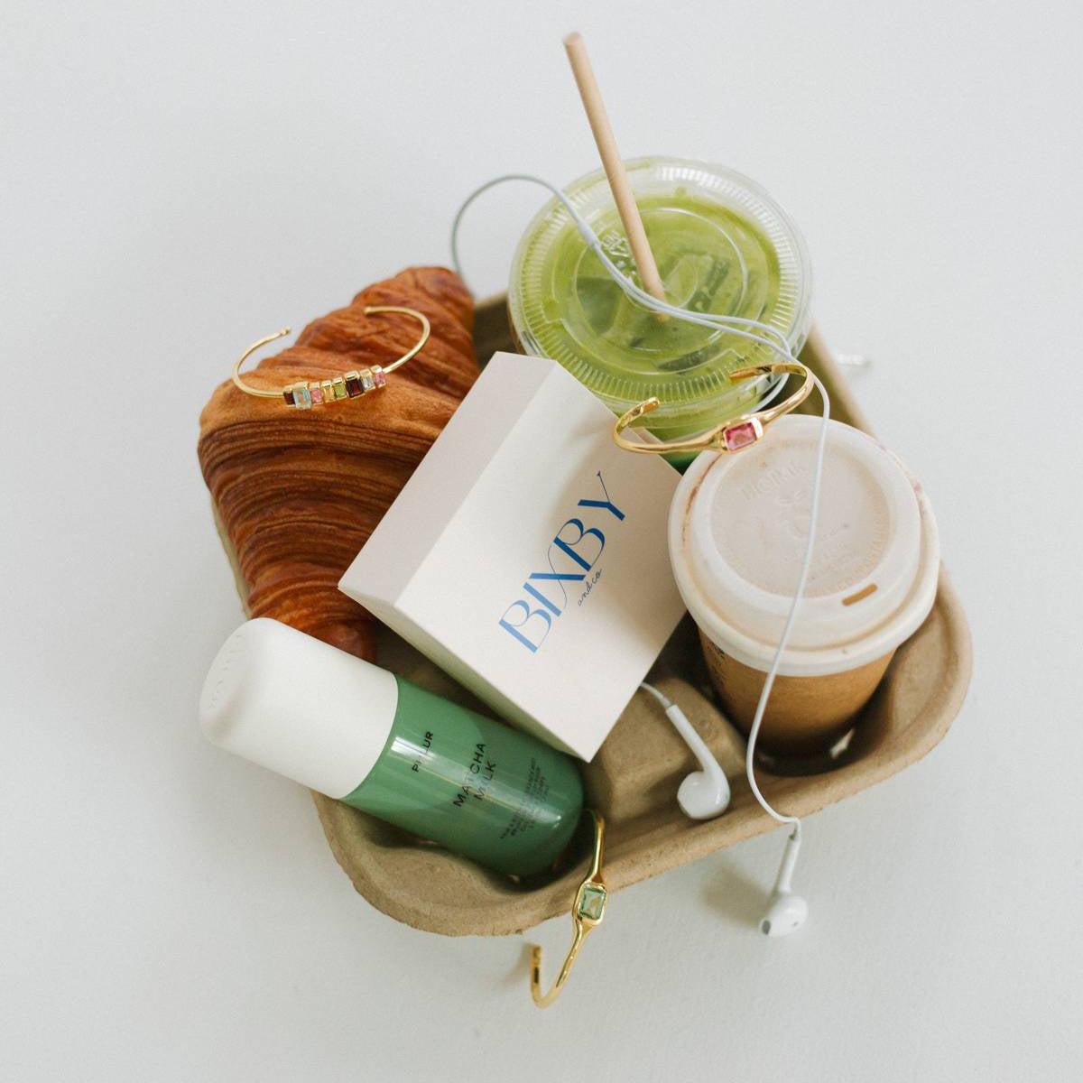 A styled flat-lay of various items arranged in a brown paper tray on a white background, including a green drink with a straw, a takeaway coffee cup, a green bottle, a white box with blue 'FANBY' branding, a croissant, and gold jewelry.