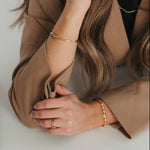 A woman in a brown blazer rests her arms on a white desk, showcasing gold jewelry including bracelets, rings, and a hoop earring. A black binder and pastel sticky notes are visible on the desk next to her.
