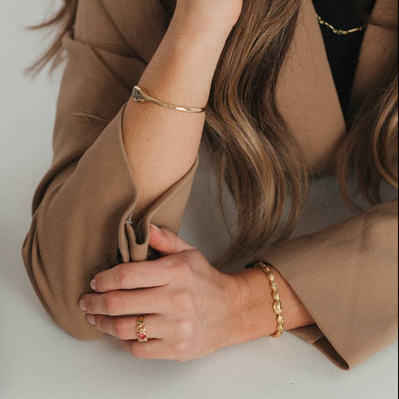 A woman in a brown blazer rests her arms on a white desk, showcasing gold jewelry including bracelets, rings, and a hoop earring. A black binder and pastel sticky notes are visible on the desk next to her.