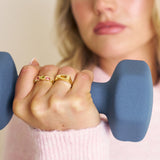 Person holding a blue dumbbell with gold rings on fingers, wearing a pink sweater.
