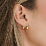 A close-up of a person's ear wearing a small gold huggie hoop earring adorned with tiny green and red gemstones.