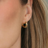 A close-up of a woman's ear wearing a small gold crescent-shaped hoop earring with a black center and tiny red and orange stones.
