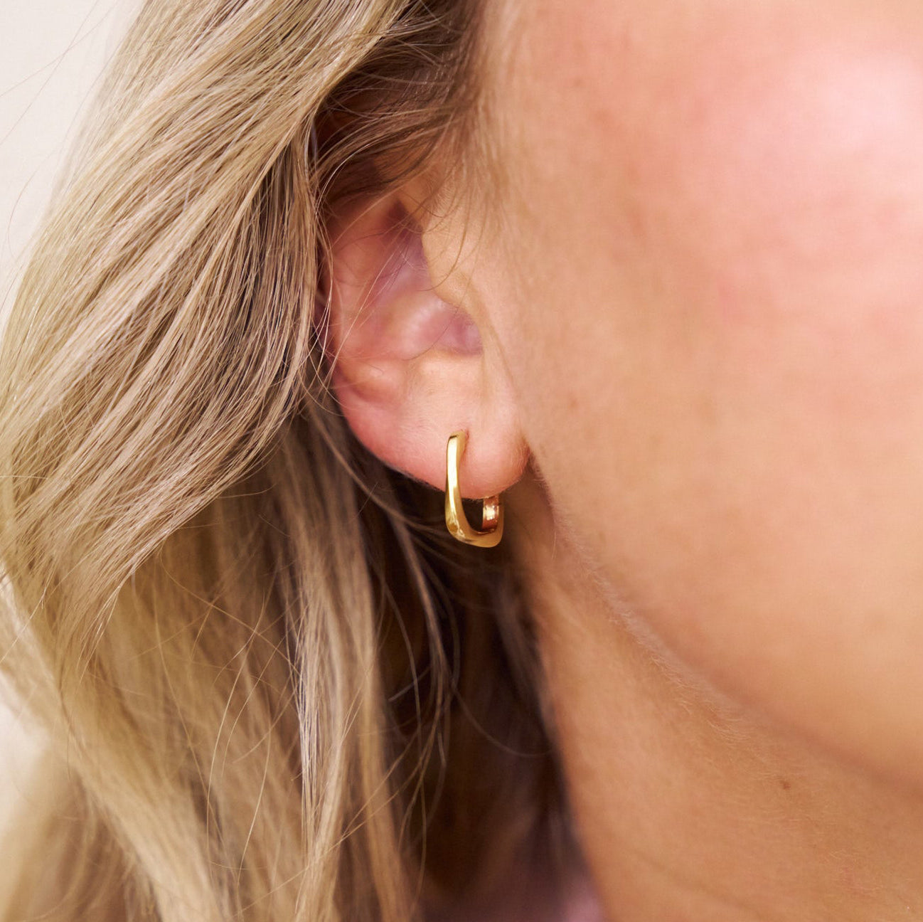 Close-up of a person wearing a square gold hoop earring with blonde hair.