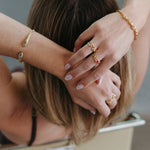 A close-up lifestyle shot of a woman from behind, showing her brown hair and hands adorned with multiple gold rings and bracelets.