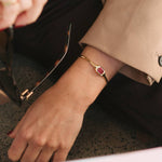 A close-up lifestyle shot of a person wearing a gold bangle bracelet with a rectangular red gemstone, paired with a beige blazer and black trousers.