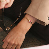 A close-up lifestyle shot of a person wearing a gold bangle bracelet with a rectangular red gemstone, paired with a beige blazer and black trousers.