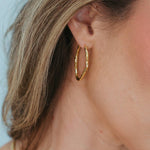 A close-up of a woman's ear wearing a textured gold hoop earring, with her hair and skin visible against a light blue background.