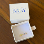 Two gold rings with light green stones displayed in an open white Bixby arka jewelry box, with the closed white lid showing the blue Bixby logo in the background, all placed on a wooden table.