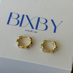 A pair of gold huggie hoop earrings with colorful rhinestone accents, displayed on a white BIXBY and Co. brand gift box on a dark wooden surface.