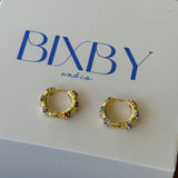 A pair of gold huggie hoop earrings with colorful rhinestone accents, displayed on a white BIXBY and Co. brand gift box on a dark wooden surface.