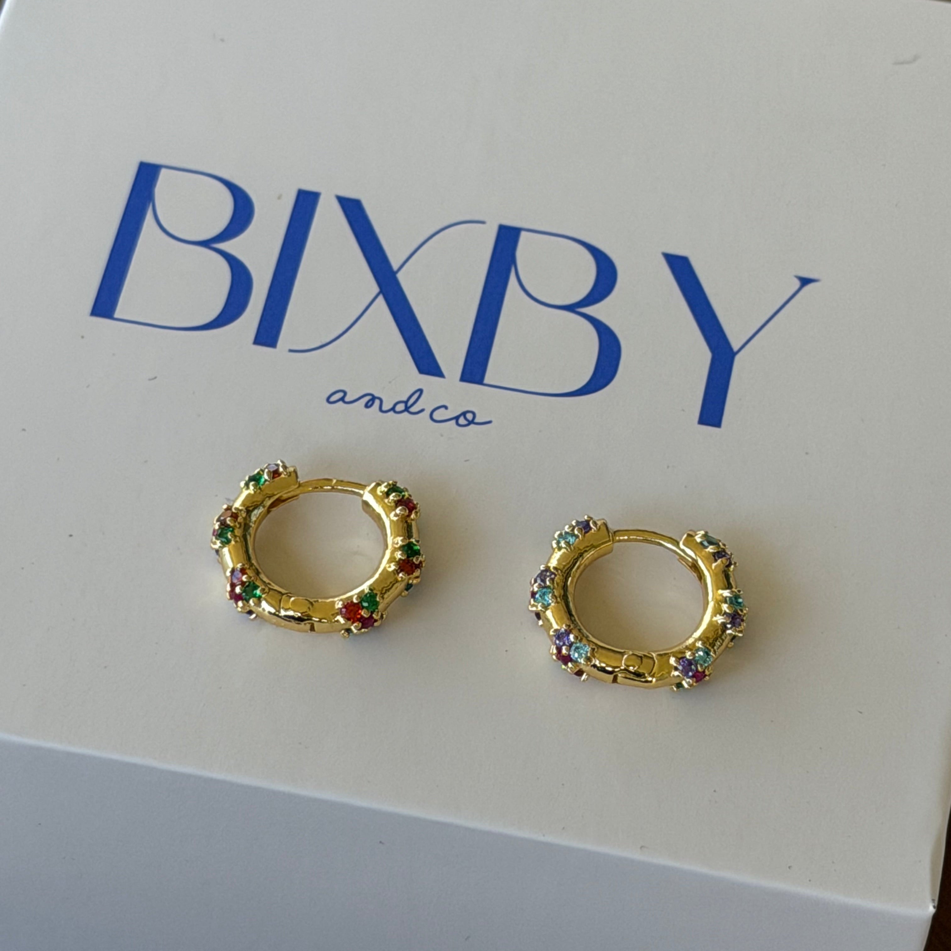 A pair of gold huggie hoop earrings with colorful rhinestone accents, displayed on a white BIXBY and Co. brand gift box on a dark wooden surface.