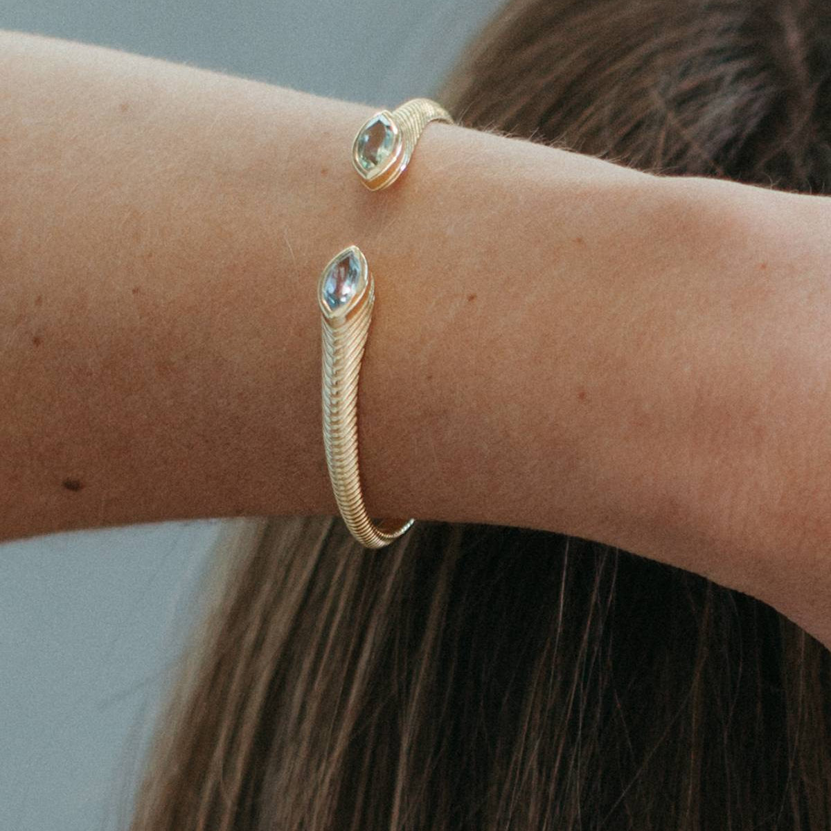 A close-up of a person's wrist wearing a gold snake-style cuff bracelet with two oval clear gemstone accents, against a soft gray background and brown hair.