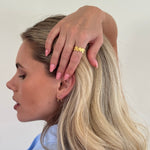 Woman with blonde hair touching her head, wearing a gold ring on a plain background