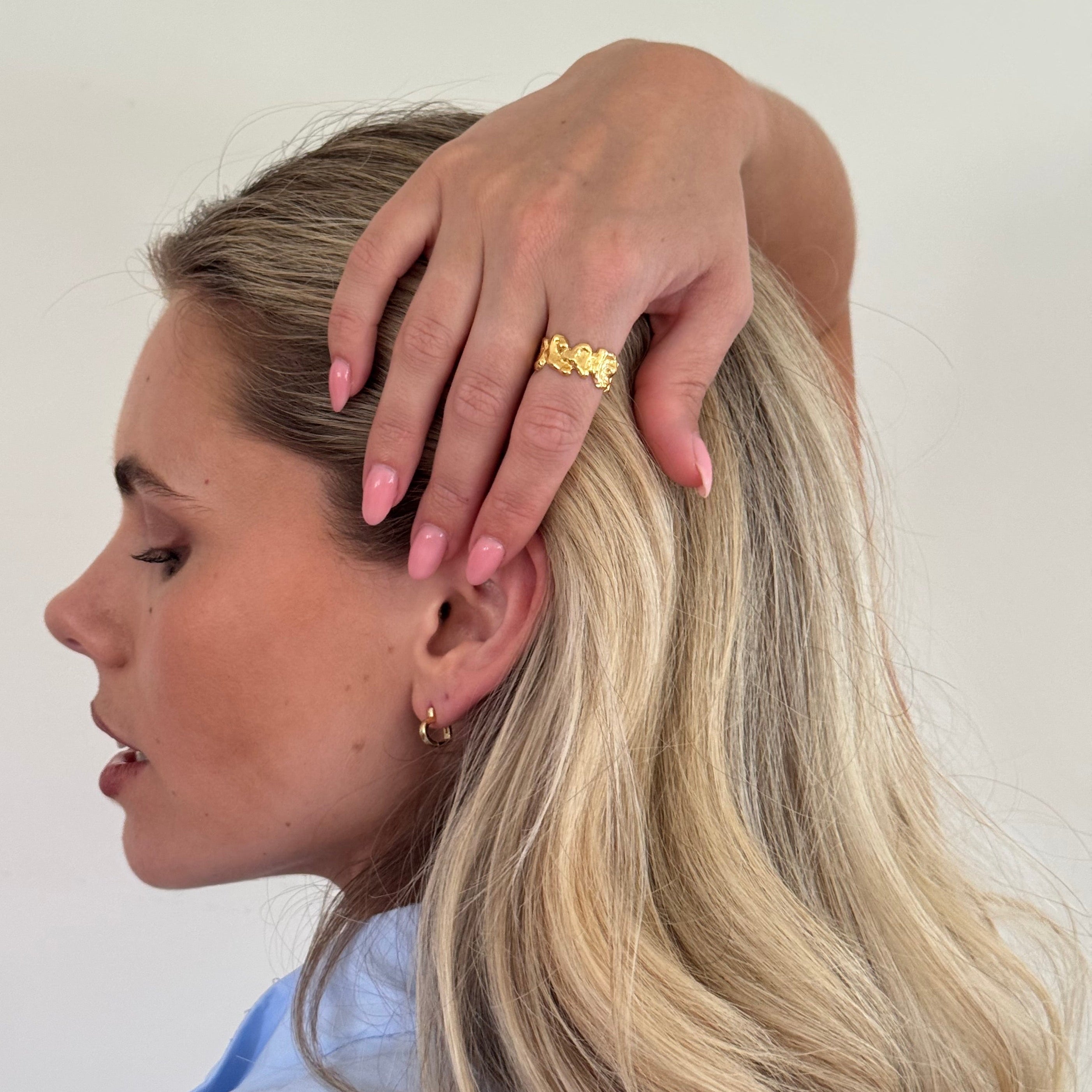 Woman with blonde hair touching her head, wearing a gold ring on a plain background