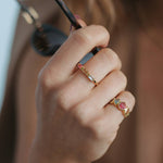 A close-up of a person's hand wearing two gold rings with colorful gemstones, along with a gold bracelet and holding sunglasses.
