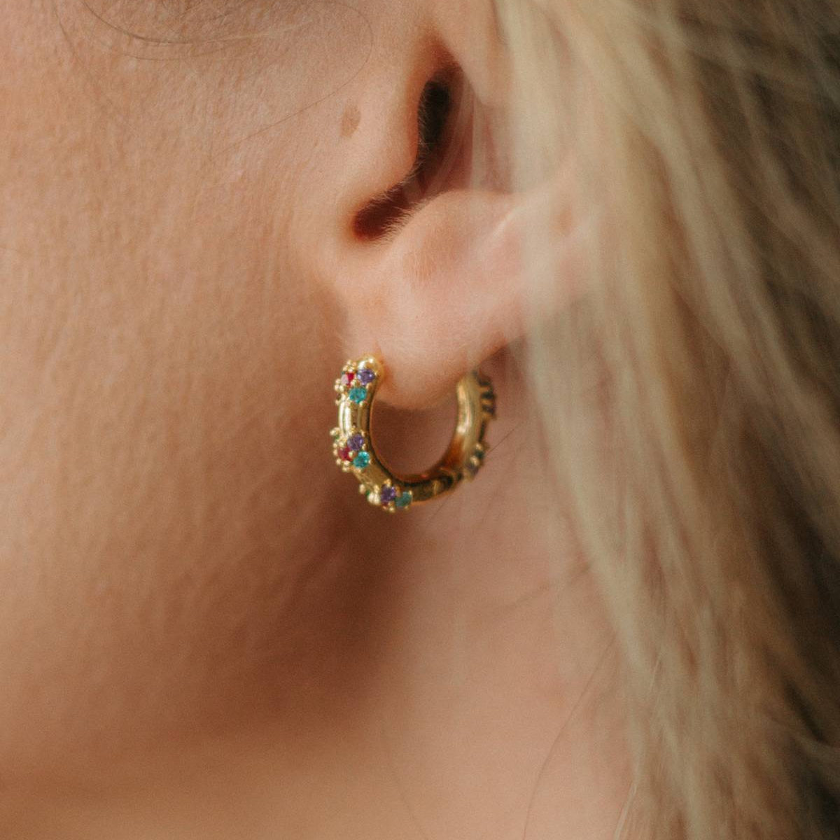 A close-up of a person's ear wearing a small gold huggie hoop earring adorned with colorful rhinestones.