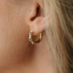 A close-up of a person's ear wearing a small gold huggie hoop earring adorned with colorful rhinestones.