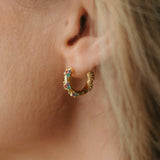 A close-up of a person's ear wearing a small gold huggie hoop earring adorned with colorful rhinestones.