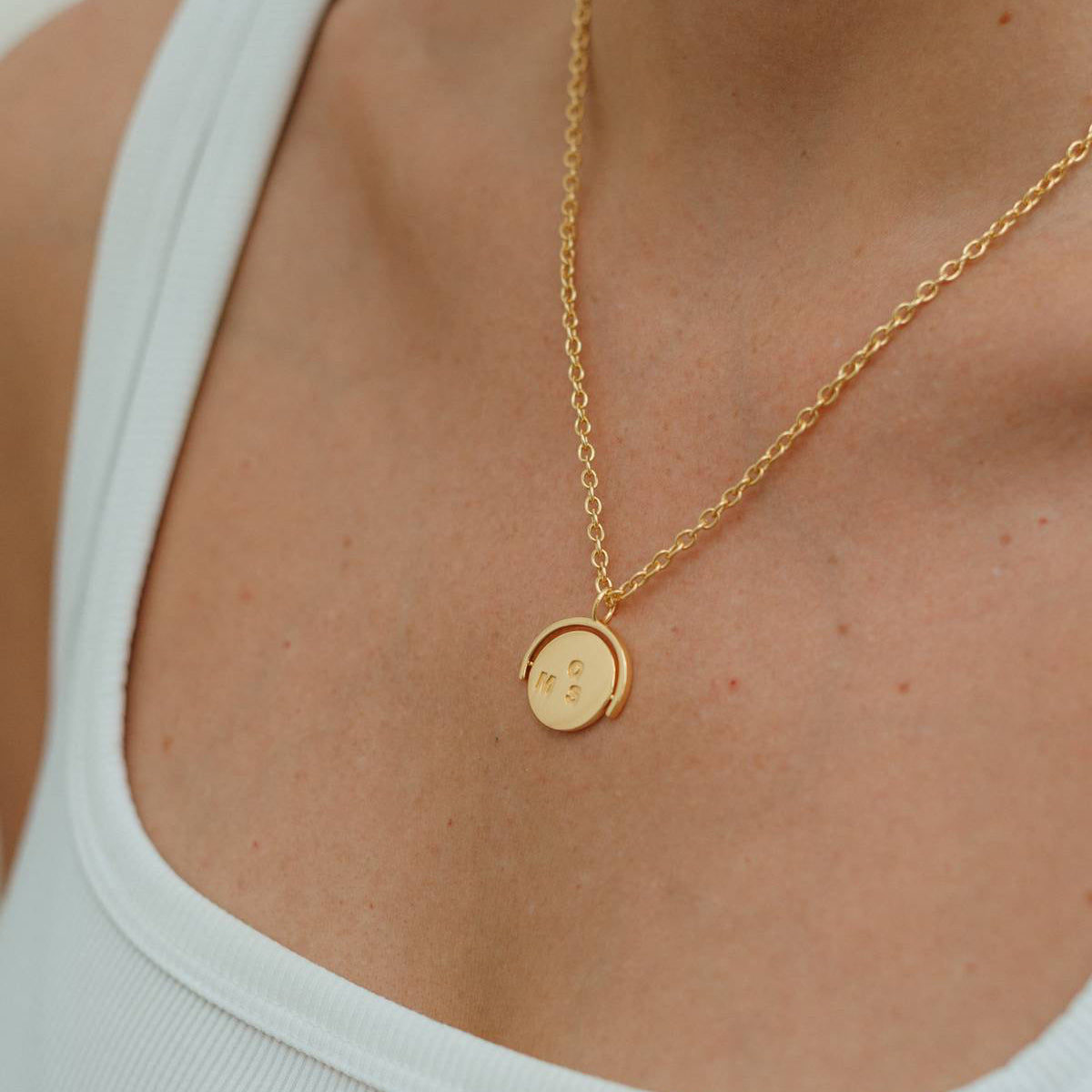 Gold necklace with a round pendant worn by a person in a white tank top.