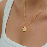 Gold necklace with a round pendant worn by a person in a white tank top.