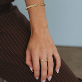 A close-up of a person's hand and wrist wearing gold jewelry, including a gold snake bracelet on the wrist and a gold double-C ring on the finger, while seated on brown pinstriped trousers against a light blue wall.