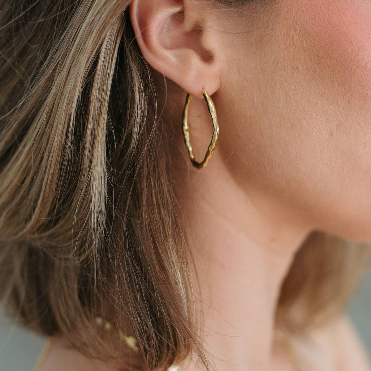 A close-up of a woman's ear wearing a textured gold hoop earring, with her brown hair and skin visible against a soft neutral background.