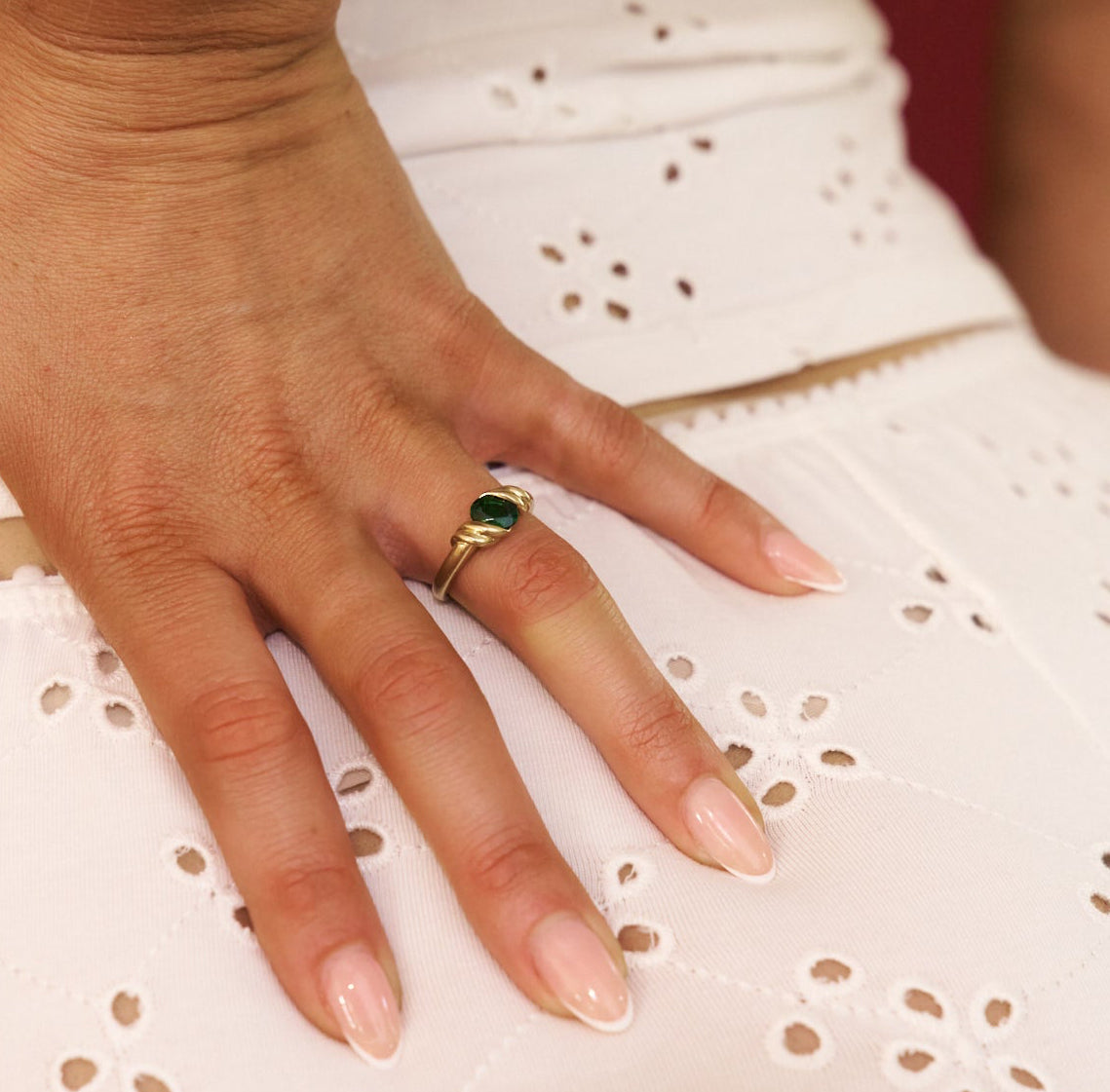 Hand wearing a gold ring with a green gemstone on a white lace fabric background