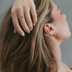 A close-up lifestyle shot of a woman with short brown hair, wearing a delicate gold ring on her finger and a small gold hoop earring, with her hand resting on her head against a soft gray background.