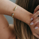 A close-up lifestyle shot of a woman's arm and hair, showcasing a delicate gold cuff bracelet with two small oval clear gemstones set at the ends.