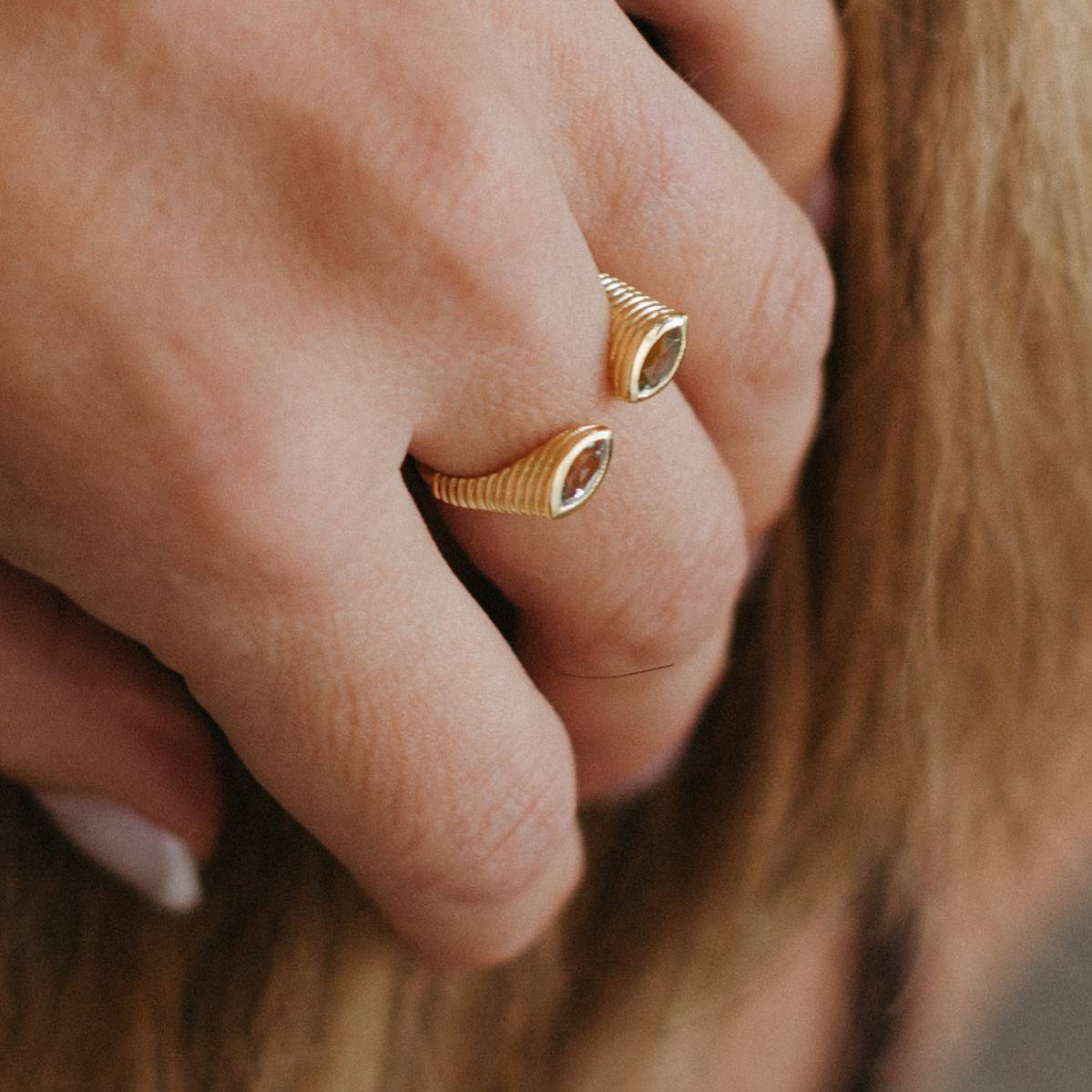 A close-up of a person's hand wearing two slim gold rings with oval clear gemstones, set against a background of light brown hair.