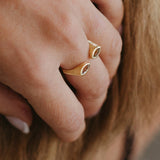 A close-up of a person's hand wearing two slim gold rings with oval clear gemstones, set against a background of light brown hair.