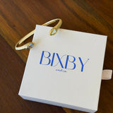 A gold snake-head cuff bracelet with clear oval gemstones resting on a white Bixby and Co. gift box, placed on a dark wooden table.