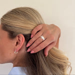 Person with blonde hair touching their head, wearing a ring and earring, on a plain background