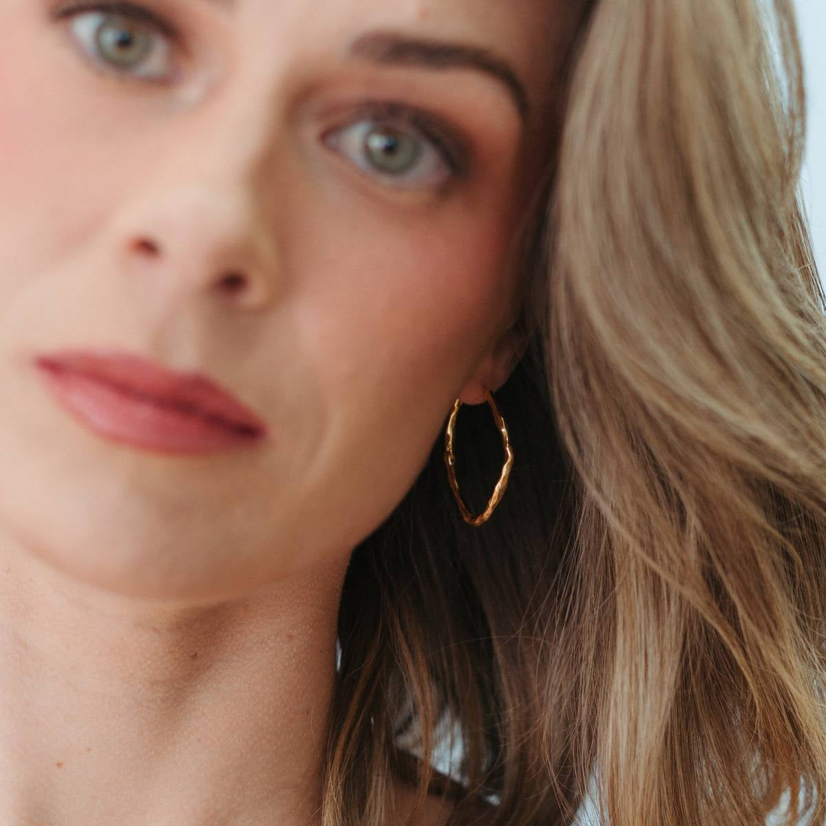 A close-up lifestyle shot of a woman with blonde hair wearing a delicate gold chain necklace and a gold hoop earring.