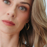 A close-up lifestyle shot of a woman with blonde hair wearing a delicate gold chain necklace and a gold hoop earring.