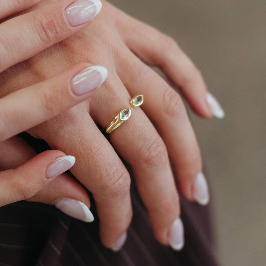 A close-up of a person's hands with manicured nails, wearing a delicate gold ring with a small oval green gemstone on one finger, set against a soft, blurred brown background.