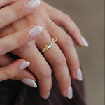 A close-up of a person's hands with manicured nails, wearing a delicate gold ring with a small oval green gemstone on one finger, set against a soft, blurred brown background.