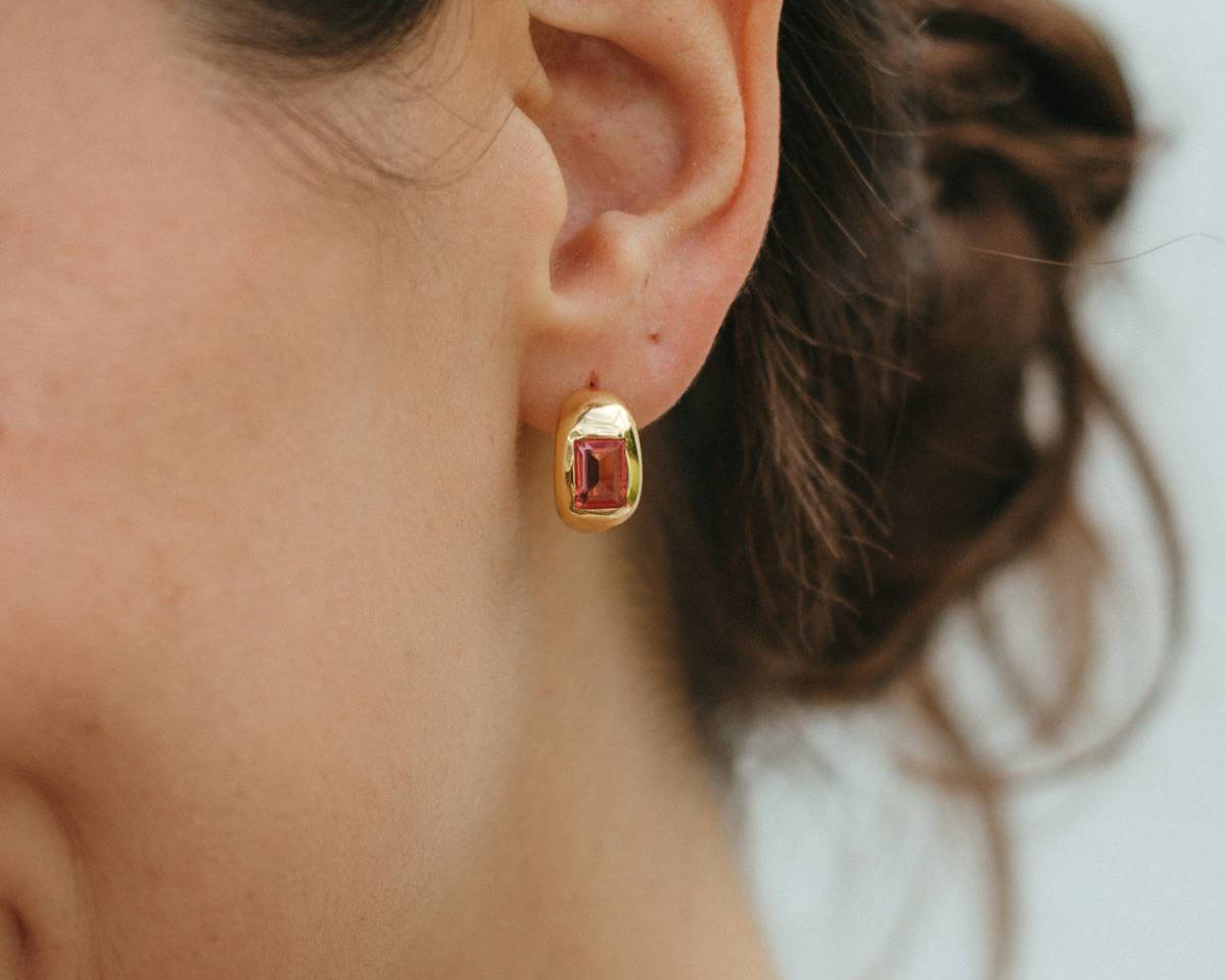 Close-up of a person wearing a gold earring with a red gemstone against a neutral background