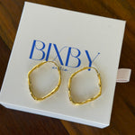 Two gold-toned, irregularly shaped hoop earrings displayed on a white BIXBY jewelry box, set against a dark wooden background.