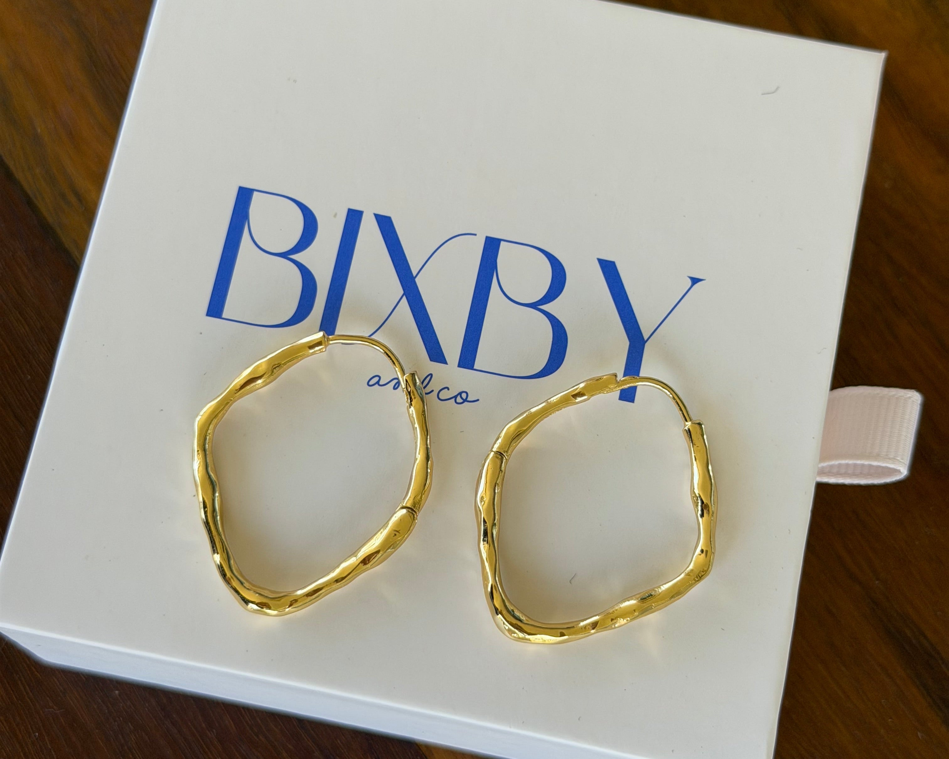Two gold-toned, irregularly shaped hoop earrings displayed on a white BIXBY jewelry box, set against a dark wooden background.