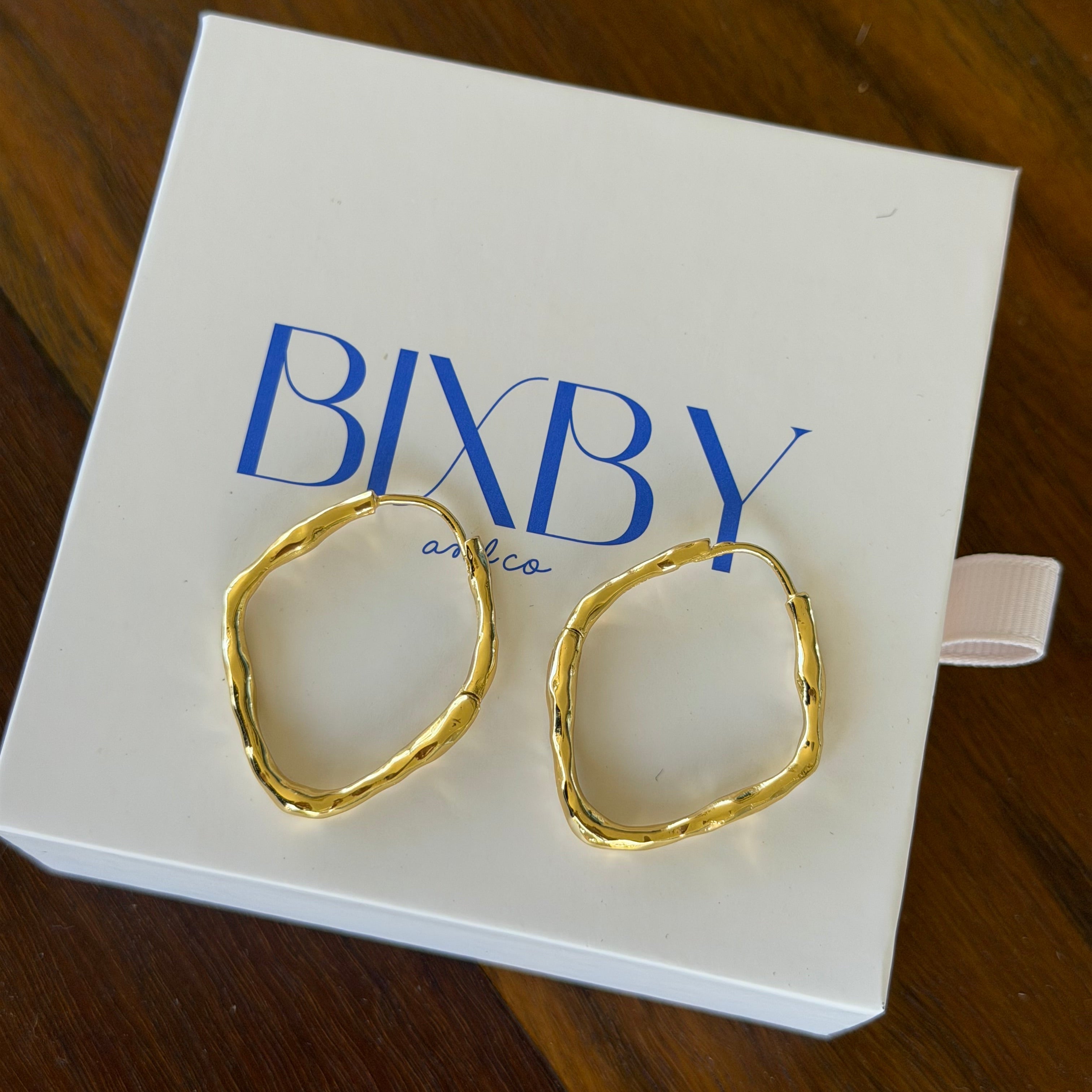 Two gold-toned, irregularly shaped hoop earrings displayed on a white BIXBY jewelry box, set against a dark wooden background.