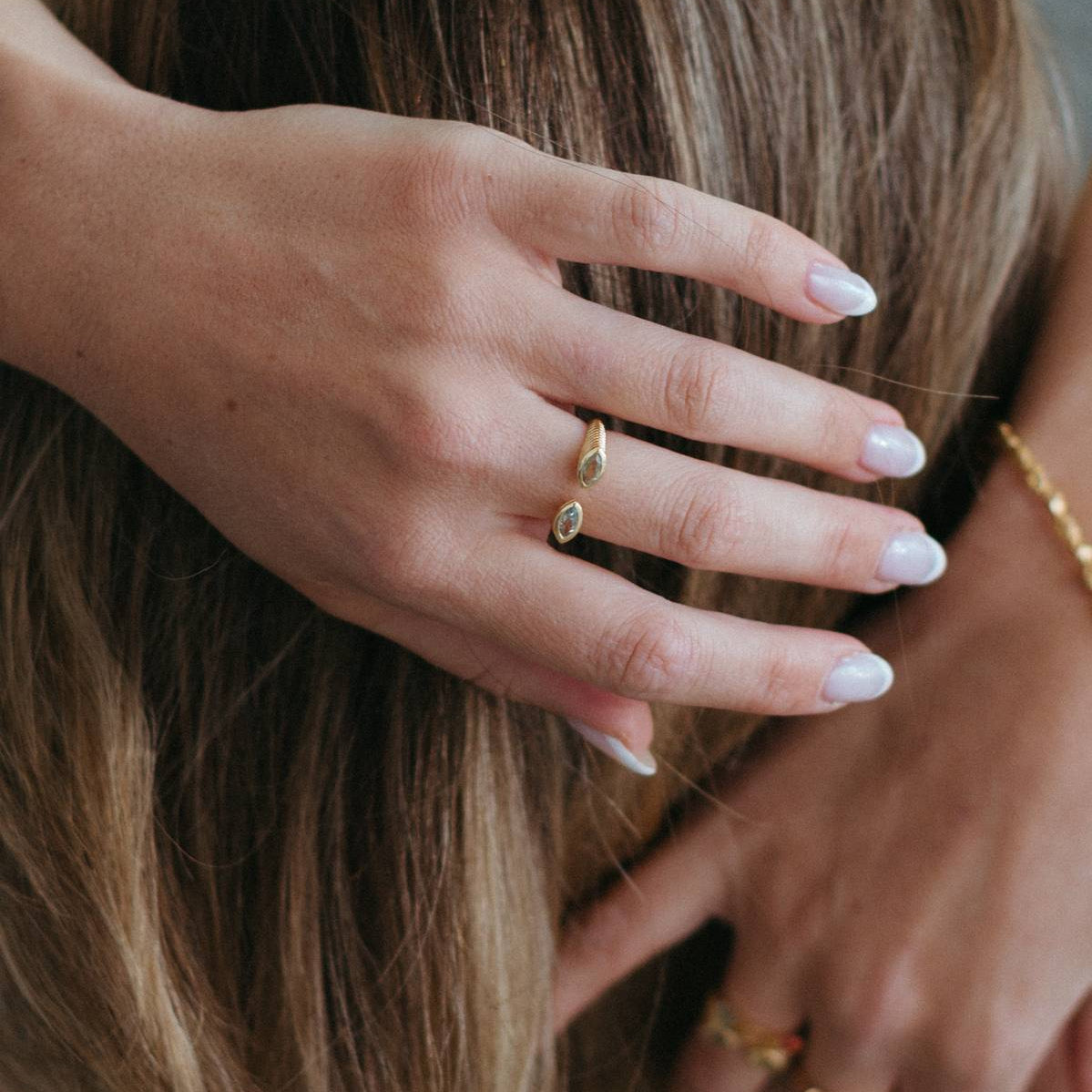 Close-up of a person with long brown hair wearing delicate gold rings, with one hand gently touching the hair. The nails are painted a light neutral shade.