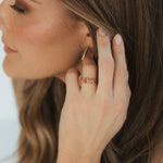 A close-up lifestyle shot of a woman wearing a delicate gold beaded bracelet on her wrist, a gold hoop earring, and a gold ring with small red gemstones, set against her brown hair and tan top.