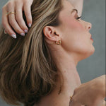 A woman with short blonde hair is shown in profile, leaning back in a chair and touching her hair and shoulder. She is wearing a dark brown ribbed camisole top and gold jewelry against a textured gray wall background.