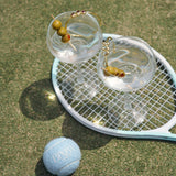 Two cocktail glasses with olives on a tennis racket on grass