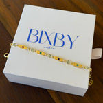 A gold-toned beaded bracelet with colorful oval beads lies across a white jewelry box printed with the brand name 'BIBY and co'. The box is placed on a dark wood surface.