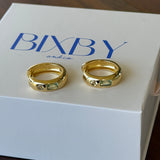 Two gold hoop rings displayed on a white BIXBY brand box, set against a dark wooden background.