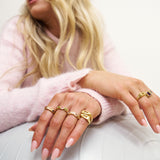 Close-up of a person's hands wearing multiple gold rings with a pink sweater and white background.