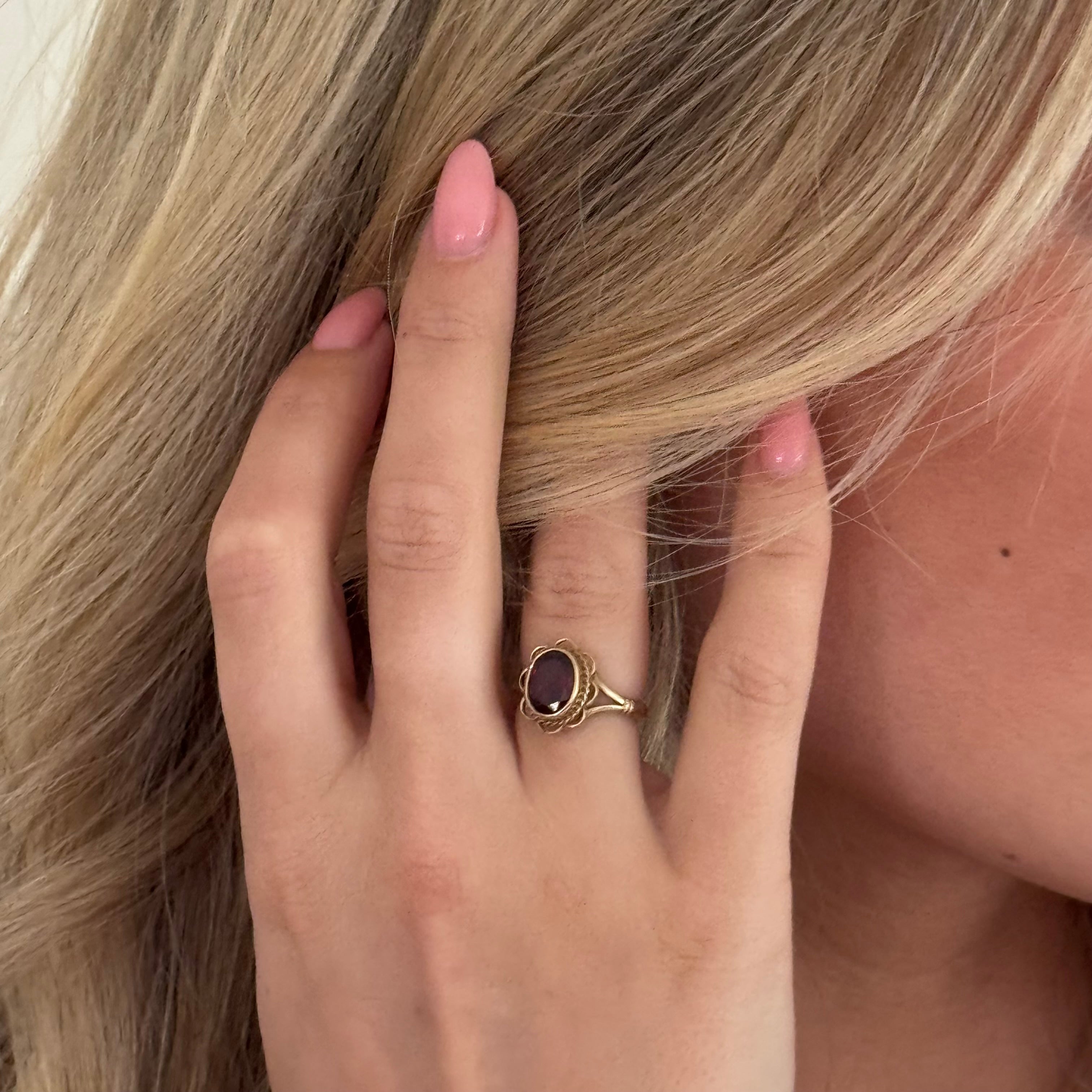 Close-up of a hand with a gold ring featuring a red gemstone, touching blonde hair.